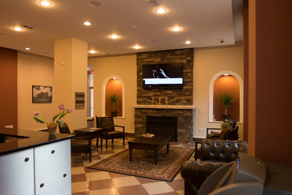 Resident lounge with a stone fireplace and a television mounted above the mantel, arched alcoves with plants on either side, and leather armchairs on a patterned rug.