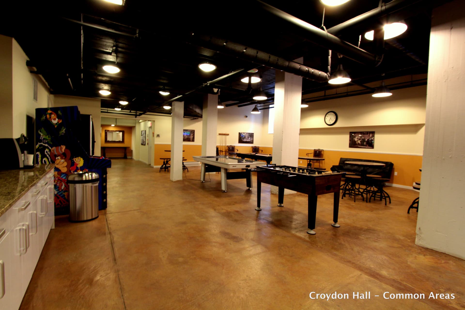 Common room with a foosball table in the foreground, an air hockey table and an arcade cabinet behind it, high-top tables and a kitchenette with white cabinets beneath a black-painted exposed ceiling.