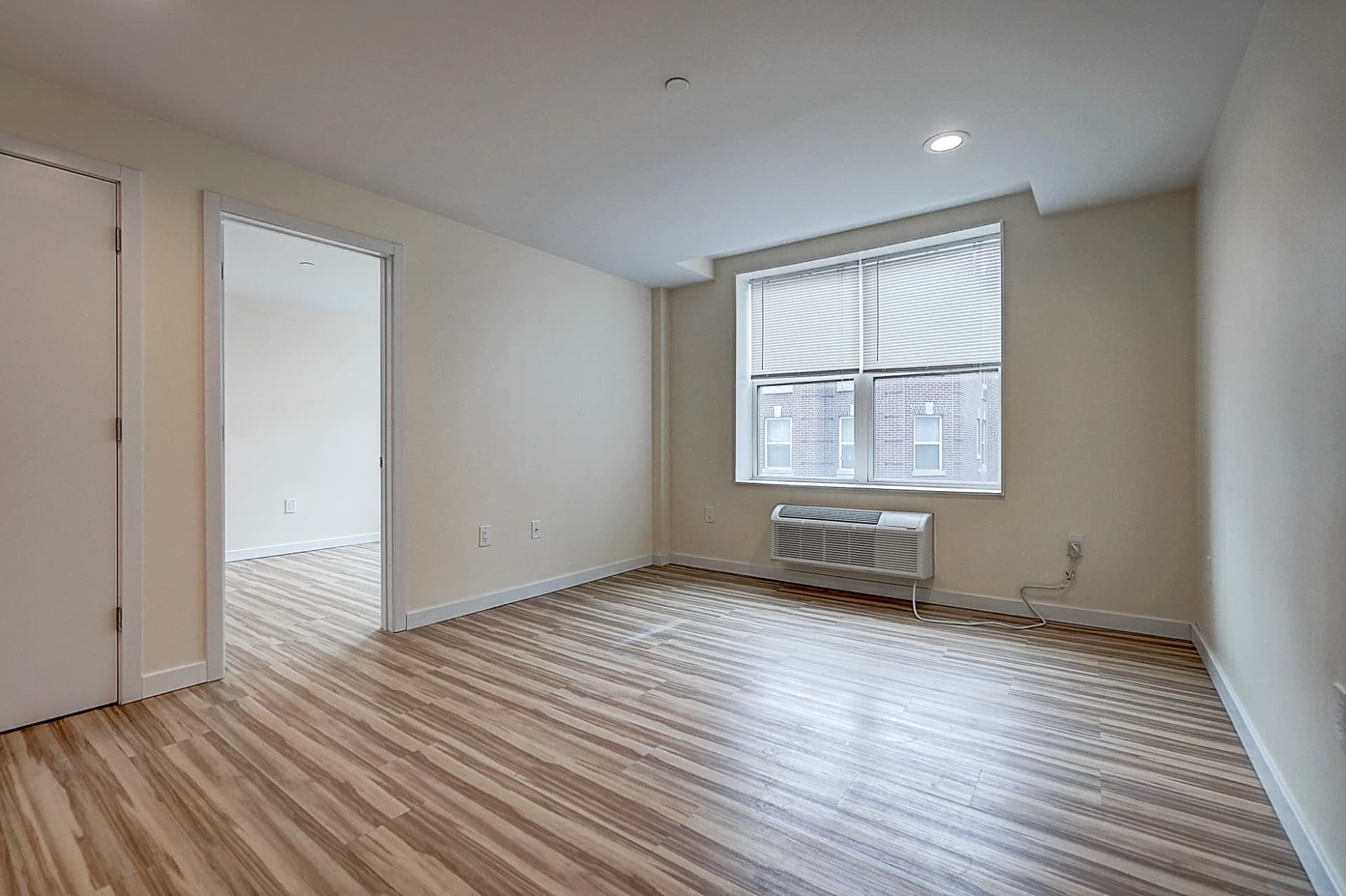 Empty living area with wood-pattern flooring, a wide window above an air conditioner and an open doorway to an adjoining room.