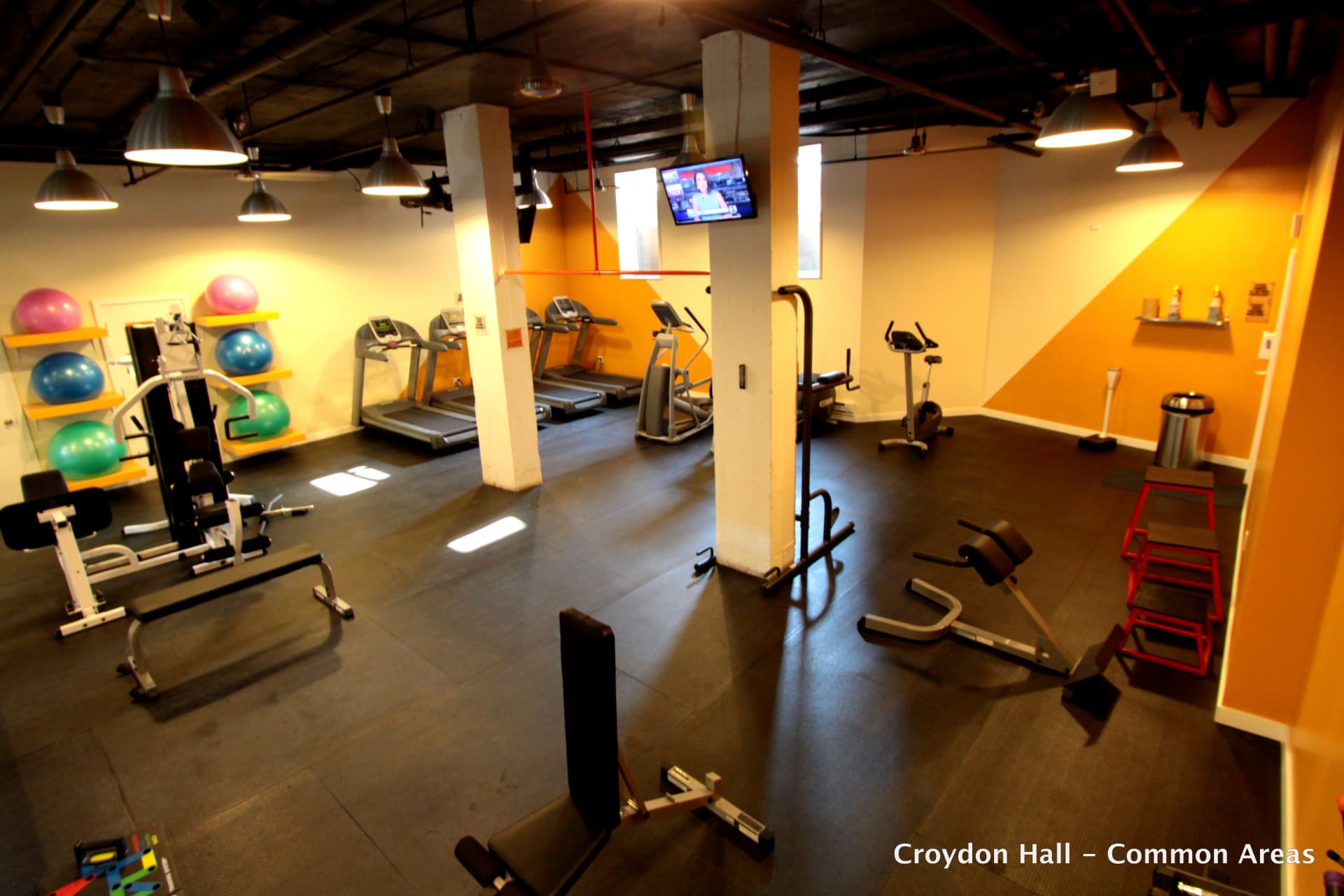 Elevated view of the fitness room showing weight benches, a cable machine, red jump boxes and an exercise bike in the foreground, with treadmills and ellipticals along an orange-accented wall.