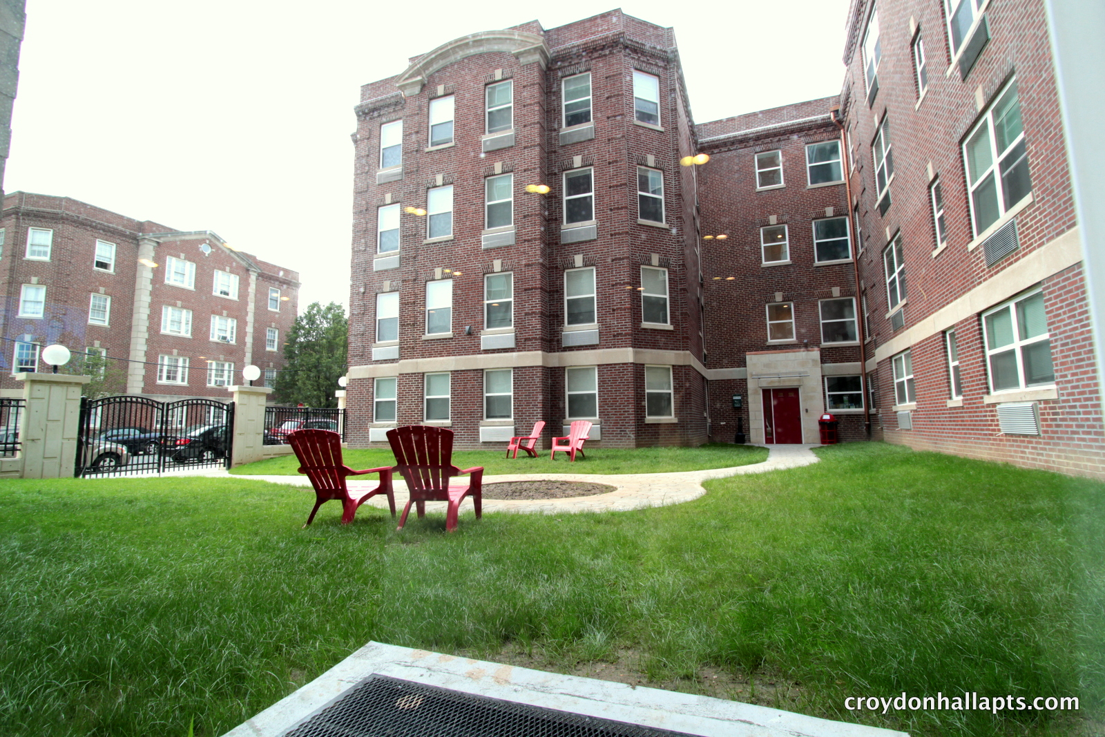Grassy courtyard surrounded by Croydon Hall's red brick apartment buildings, with red Adirondack chairs beside a curved walkway and a black metal gate on the left.
