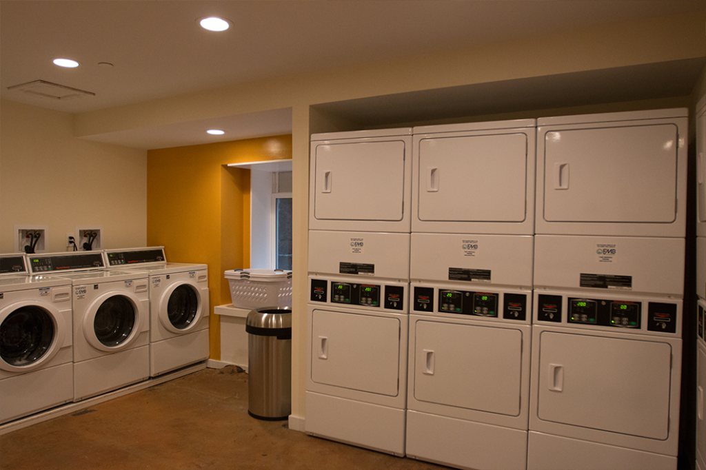 On-site laundry room with three front-loading washers along one wall and stacked dryers recessed along the other, lit by recessed ceiling lights.