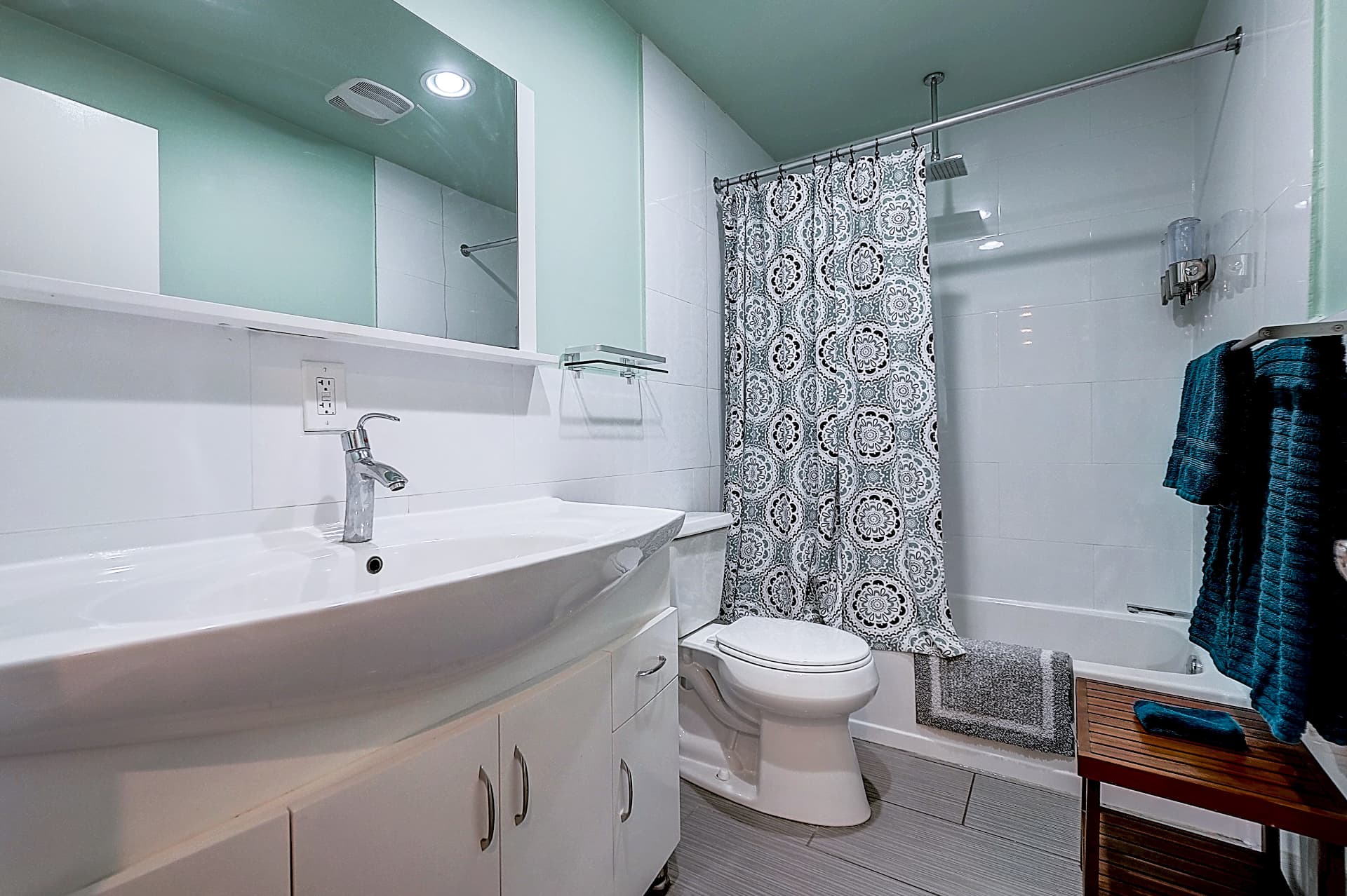 Furnished bathroom with a wide white vanity and mirror, a toilet, patterned shower curtain, blue towels and a wooden bath mat.