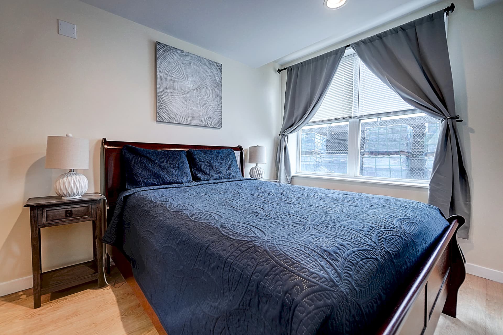 Furnished bedroom with a dark wood bed, blue quilt, two bedside lamps and gray curtains framing a large window.