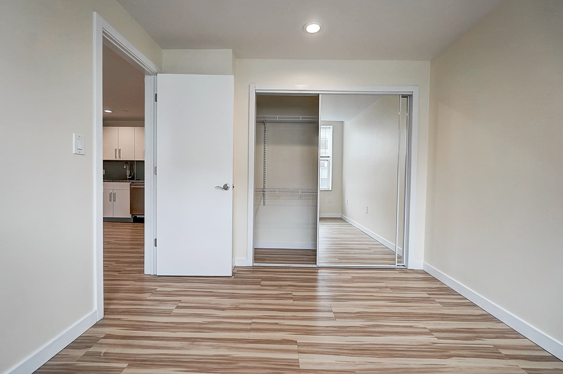 Unfurnished room with wood-pattern flooring and a partially open mirrored sliding closet showing wire shelves; a doorway leads toward the kitchen.