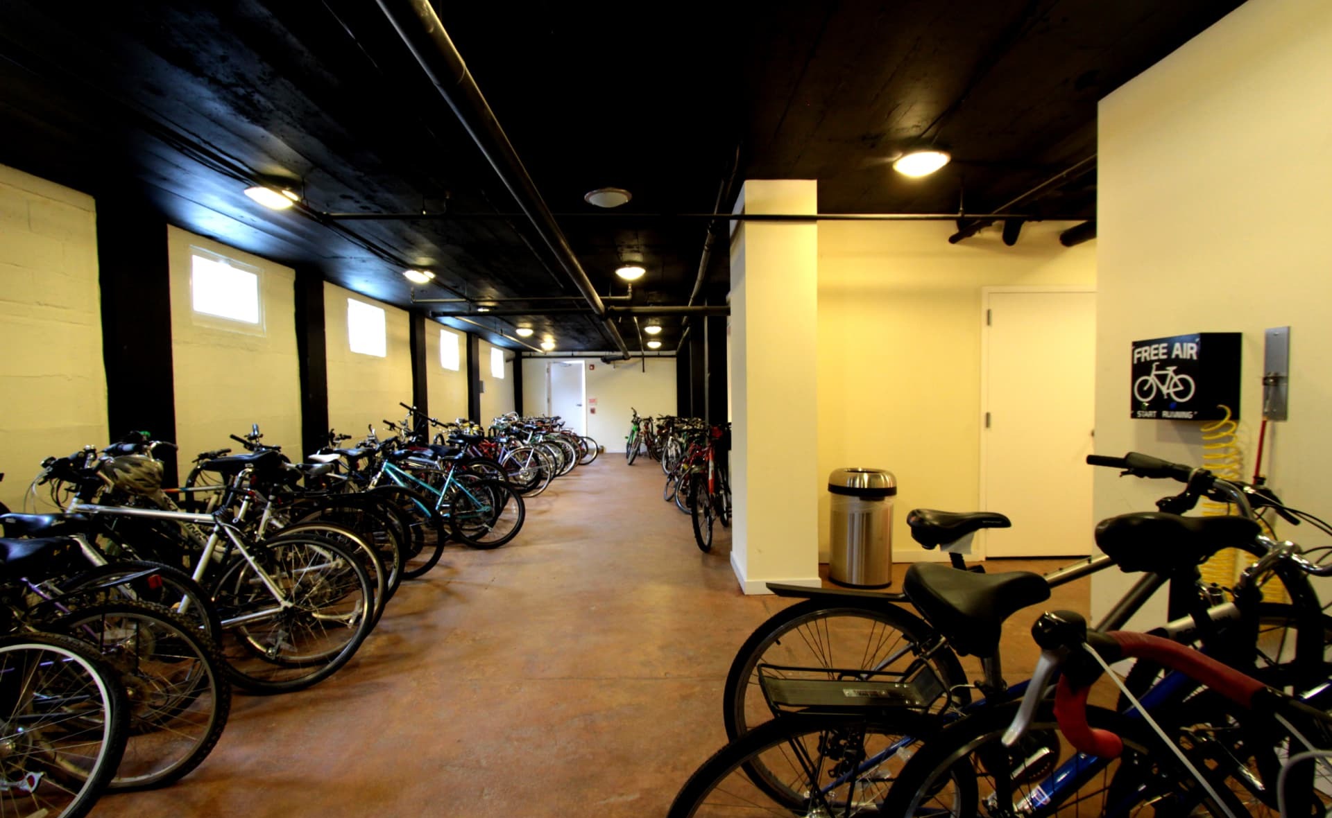 Indoor bicycle storage room with bikes parked along both walls and a wall-mounted free air pump station with a coiled hose, lit by high windows and warm ceiling lights.