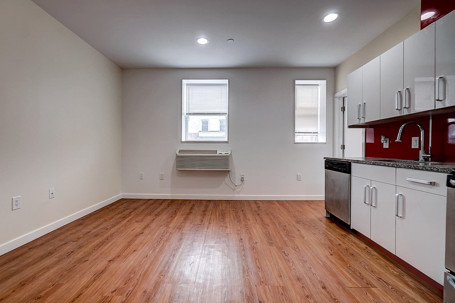 Empty living area with wood flooring, two windows and a wall-mounted air conditioner; white kitchen cabinets line the right wall.