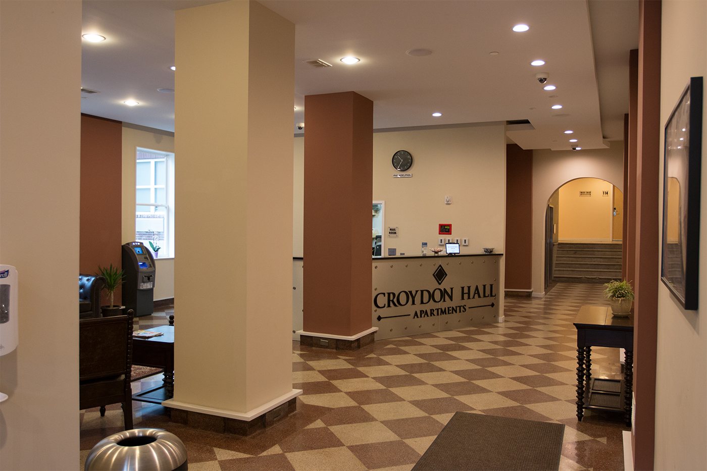 Croydon Hall Apartments lobby with a curved metal reception desk bearing the building's name, leather seating and an ATM to the left, and a checkered tile floor.