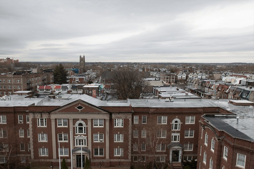 Rooftop view across the brick buildings and row houses of West Philadelphia, with a stone church tower on the horizon under a gray winter sky.